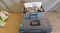 Closed black plastic toolbox containing miscellaneous tools with a handle on top, placed next to a blue box filled with various tools including squares, chisels, files, and masonry tools.