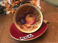 Teacup placed inside saucer showing interior fruit motif and exterior red glaze with gold trim