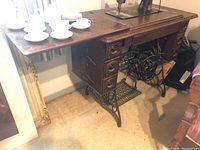 Full view of antique Singer treadle sewing machine with wooden surface and decorative carved drawers on sides. White ceramic cups placed on the base as scale reference.
