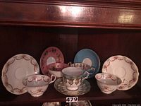 Five Aynsley porcelain teacups and saucers arranged on a wood shelf, showing varied floral and gold trim patterns.