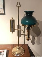 Full view of brass oil lamp with green glass shade and brass cylindrical component on the side, standing on a wood surface.
