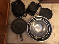 Image of cast iron cookware and stainless steel bowls on tiled floor. Includes frying pans, muffin pan, colander bowl, and enamel roasting pan.