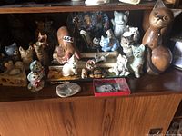 Shelf with multiple cat figurines in various poses, styles, and materials, along with cat-themed books beneath them.