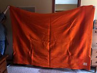 Full view of orange wool blanket held horizontally by two people against a living room background