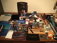 Wide view of desk with multiple office supplies and accessories including calculators, clocks, pens, and novelty pen holders