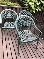Three green metal patio chairs with lattice design on the seat and backrest shown on a wooden deck next to a fence with ivy.