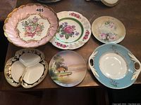 Overview photo showing six porcelain serving pieces including a pink cake stand, several floral plates, and a divided serving dish.