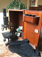 Full view of vintage microscope and open wooden case showing accessory compartments with lenses and slides, black cloth on ground