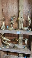 Nine brass figurines displayed on wooden shelves featuring various bird species and dolphins. Includes cranes, swans, and dolphins in multiple poses and sizes.