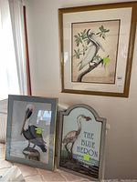 Three items: two framed pelican artworks and a Blue Heron wall plaque. The pelican artworks show pelicans perched on branches with soft natural backgrounds, framed in brown and gold frames. The plaque is a decorative piece with a stylized blue heron and text.