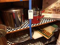 Wide view of two wooden shelves holding kitchen items including large stainless steel pots and pans, smaller red pots, boxed glassware tumblers, cutting boards, and metal tray.
