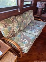 Vintage rattan three-piece sofa with six cushions featuring a tropical leaf pattern, shown in ambient natural light on wooden floor near windows.