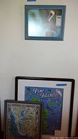 Three framed wall art pieces hung on a wall: top is a framed pelican print, below are two framed maps including puzzle and illustrated map.