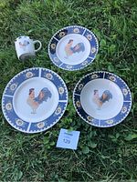 Dinnerware items including one mug, two salad plates or dessert plates, and one bowl, all with a rooster in the center and a blue border with sunflower patterns, shown on grass.