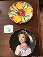 Top and front view of the colorful flower plate and the Queen Elizabeth II Silver Jubilee plate on a wooden surface with a lot number.