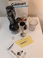 Full view of the Cuisinart Smart Stick hand blender components and box, showing the motor body, chopper container with lid, whisk attachment, measuring cup, and manuals.