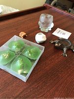 Photo of frog decor items including four green floating frog candles in original box, tiny wooden frog, white ceramic toothpick holder, clear glass frog figurine, and bronze-tone frog figurine on wooden surface.
