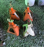 Four metal jack stands placed on gravel ground; three orange and one white, all showing signs of wear and rust.