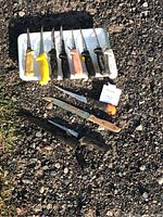 View of 11 various butcher knives laid outdoors on gravel and a white tray. Knives have different handle colors including black, yellow, white, pink, and wood. One has an orange safety cap on the blade tip. Some knives have sheaths, others don't.