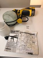 Full view of the paint sprayer laid on table with attached container, black cable, yellow trigger, and instruction manual.