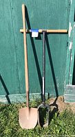 Full view of post hole digger and spade shovel against a green wooden wall on grass.
