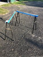 Two foldable metal sawhorses standing on an asphalt surface outdoors showing blue top bars and black legs