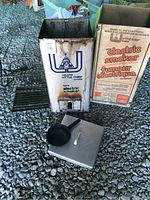 Main metal electric smoker unit with visible rust, front door, vent and a black rack beside it, along with a small pan and a thermometer. The original cardboard box with branding and model visible is behind the item.