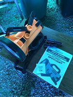 Full view of orange and black DC powered spot light with power adapter and instruction manual on wooden surface, shot outdoors with natural light.