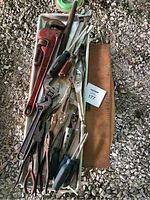 Overview of assorted hand tools arranged on a white plastic tray on gravel ground. Includes a red pipe wrench, screwdrivers, pliers, and adjustable wrenches.
