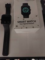 Black silicone smart watch alongside its original packaging box showing the watch face display and branding.