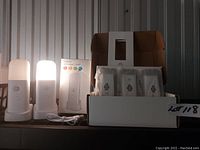 Photo showing two LED night lights turned on with frosted covers, USB charging cable, four small packaged items, and an open box containing the items.