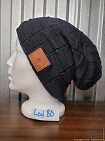 Side view of black quilted knit toque with leather patch showing Bluetooth speaker control buttons.