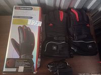 Two SabotHeat battery heated gloves in black with red accents and adjustable wrist straps, displayed next to original packaging and charging cable.