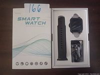 Front of the product box showing Smart Watch text, wave design, and icons indicating features. Box contains the smart watch, black silicone wristband, and USB charging cable.