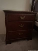 Clear image of the cherry wood nightstand showing three drawers and brass handles, with visible scratches on the surface.