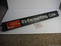 Full view of the black metal sign with white text "It's the real thing. Coke." and Coca-Cola logo on left side.