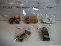 Five vintage Hot Wheels toy cars in individual plastic bags labeled with their years and models. Cars include red 4x4, yellow and blue race car, beige van, white sports car, and black sedan from the 1980s.