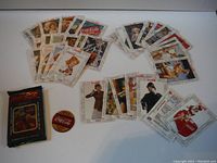 Photo showing the collection of vintage Coca-Cola collectible cards arranged in fan shapes on a white surface, with the empty original packet and a round promotional pin/button nearby.