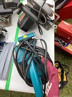 Photo showing Makita belt sander in blue color with dust bag and cord, placed on a tabletop with other items.