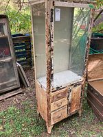 Front angled view of weathered steel display cabinet with glass door, three drawers, and solid door. Shows ongoing paint peeling and rust.