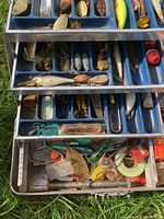 Open metal tackle box showing multiple trays filled with assorted fishing lures, spoons, hooks, and some bobbers. Various colors and types of lures visible including crankbaits, spoons, and spinners.
