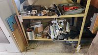 Hydraulic car jack in gray tray with other tools and tubes of caulking stored underneath a shelf.