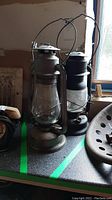 Two weathered metal Beacon hurricane lanterns with wire handles and glass chimneys on a surface.