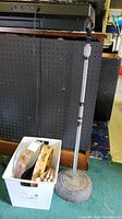 Full view of microphone stand upright next to a container holding two tambourines and wooden drumsticks on carpet.