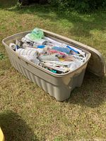Wide view of beige plastic bin open on grass filled with assorted paint supplies including rollers, brushes, sanding blocks, and tools.