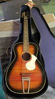 Complete guitar in open hard shell case showing sunburst body, white pickguard, trapeze tailpiece, loose steel strings and open-gear tuners