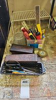 Photo showing assortment of hand tools including scissors, various screwdriver types, box cutter, wooden whetstone boxes, and wire brush attachment on workbench.