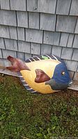 Side view of the large wooden fish painted in yellow and blue with brown fins, resting against grey wood siding on grass.