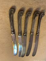 Top view of five antique sterling silver butter knives with scroll-design handles arranged side by side.