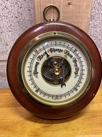 Front view of antique weather meter showing mahogany wood frame, brass rim, dial with weather condition markings, and visible mechanical components.